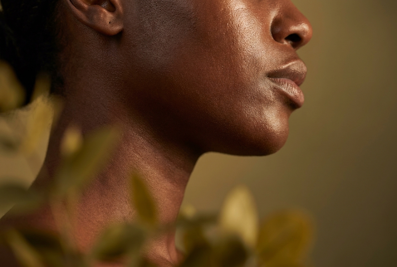 Close-up profile of a woman's jawline with deep mahogany skin, highlighting a healthy, naturally radiant complexion.